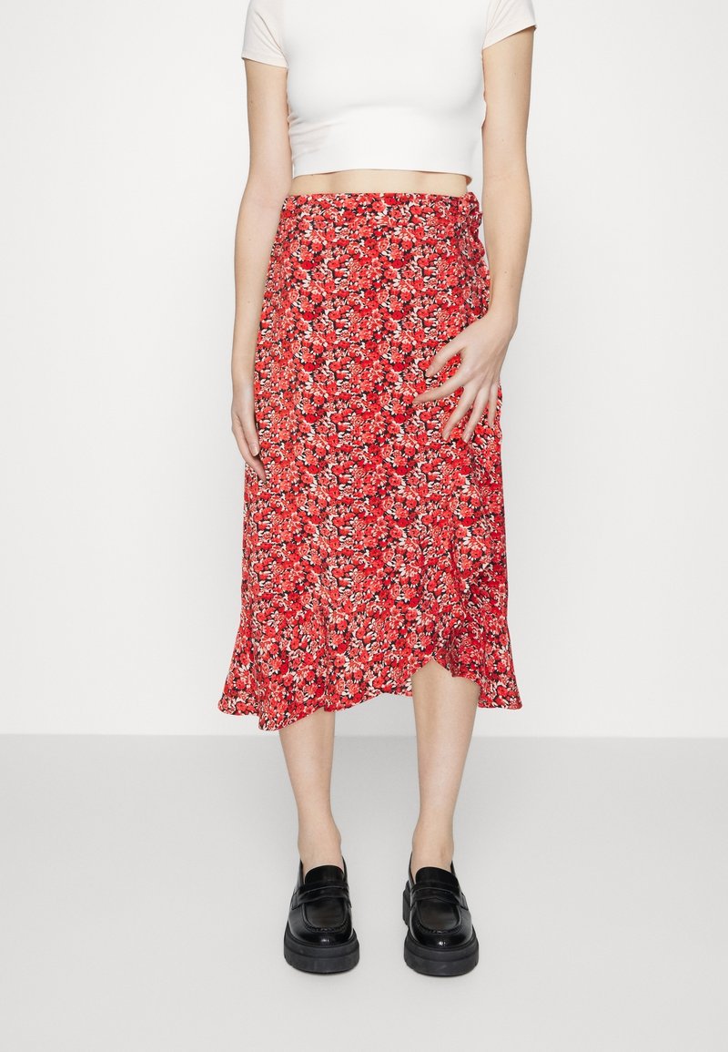 Jupe midi rouge à fleurs avec un ourlet volanté et un tissu léger, associée à des mocassins noirs et un top court blanc.