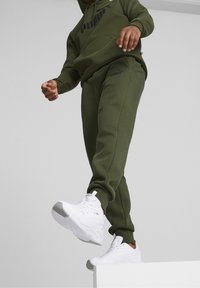 Olivgrön hoodie och sweatpants, med en framficka och logotyp. Vita sportskor med grå accenter och strukturerade sulor.