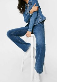 Modrá džínová bunda a rozšířené džínsy s kontrastním prošitím, výraznými kapsami a ošoupanou texturou, doplněné o bílé tenisky.