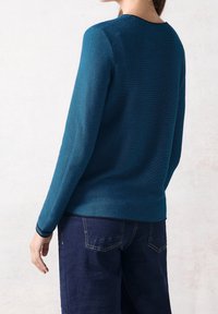 Sweter z długim rękawem w gradientowym odcieniu teal, z poziomą fakturą prążkowaną, okrągłym dekoltem i gładkimi granatowymi krawędziami. Połączony z ciemnymi dżinsami.