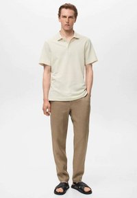 Polo shirt scurt cu mânecă în nuanță bej deschis, asociat cu pantaloni maro și sandale negre. Cămașa are un guler clasic și o textură netedă.