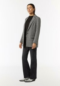 Grau strukturierter Blazer mit Reverskragen, einreihigem Knopfverschluss und seitlichen Taschen, kombiniert mit einem schwarzen Top und schwarzen Schlagjeans. Silberne Absatzstiefel.