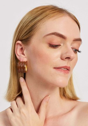 Hnedé živicové náušnice v podobe kruhu s textúrovaným povrchom a zlatým kovovým prvkom, nosené na uchu osoby, ktorá si jemne dotýka krku.