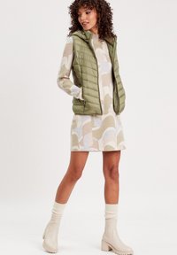 Chaleco acolchado verde oliva con capucha, usado sobre un vestido estampado en beige, azul y marrón. La modelo lleva botas de tobillo beige y calcetines crema.
