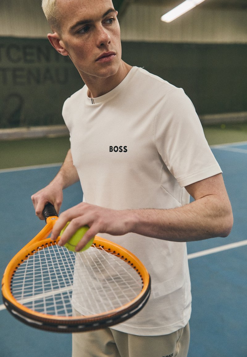 Valge sportlik särk, millel on "BOSS" logo, beežid püksid, hoiab tennisereketit, millel on oranž raam ja roheline pall, sinisel tennisekeskkonnal.