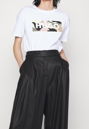 Persona con camiseta blanca con logo "HUGO" de camuflaje multicolor y pantalones negros de pierna ancha y talle alto con las manos en los bolsillos.