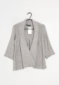 Cárdigan grueso de punto gris con un amplio cuello tipo chal, mangas cortas y patrones de diamante a los lados. Cuelga de un gancho negro.