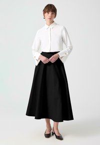 Camisa blanca de botones con mangas largas y adornos dorados, combinada con una falda negra de línea A de cintura alta y zapatos de tacón cuadrado marrones.