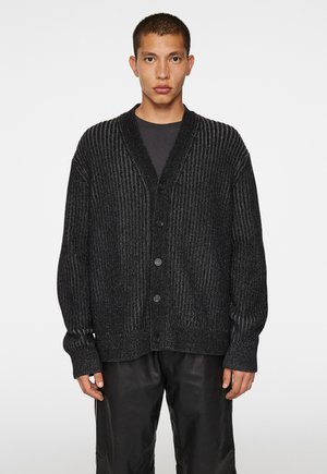 Gilet noir à côtes avec un col châle, possédant un devant à boutons et des manches longues. Porté par-dessus une chemise sombre et assorti avec un pantalon noir.