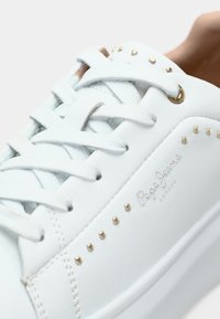 Λευκά αθλητικά παπούτσια από λείο δέρμα με χρυσές λεπτομέρειες, που διαθέτουν απλό σύστημα κορδονιών, μικρές χρυσές καρφίτσες και ανάγλυφο λογότυπο.