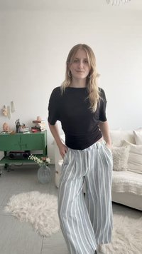 Nuori nainen, jolla on pitkät vaaleat hiukset, musta toppi ja raidalliset leveälahkeiset housut, seisoo viihtyisässä olohuoneessa vihreän kaapin vieressä.