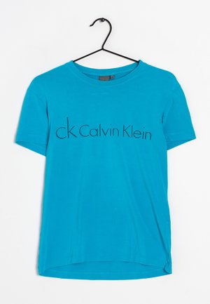 Maglietta a maniche corte di colore blu brillante con il logo "ck Calvin Klein" stampato sul petto, appesa a una gruccia di filo nero su uno sfondo bianco.