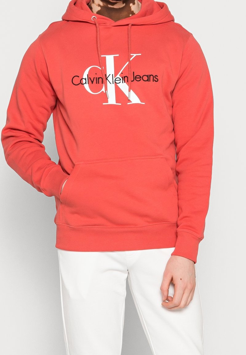 Homme portant un sweat à capuche Calvin Klein Jeans rouge corail avec logo blanc, assorti à un pantalon blanc, debout avec une main dans la poche.