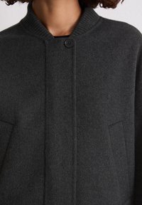 Donkergrijze wollen jas met een ribbelkraag, enkele knoopsluiting en twee zijzakken. Zachte textuur met strakke lijnen en een minimalistisch ontwerp.