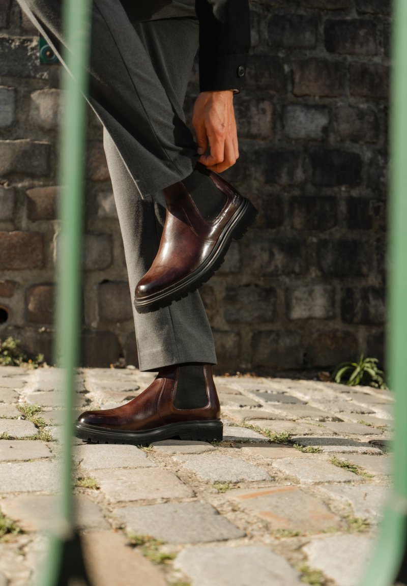 Botas chelsea de cuero marrón con paneles elásticos en los lados y suelas texturizadas, combinadas con pantalones grises, sobre un fondo de pavimento de piedra.
