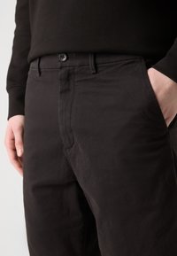 Pantalon en coton noir avec une texture lisse, doté d'une fermeture à bouton et de multiples poches, présenté de la taille jusqu'au niveau des hanches.