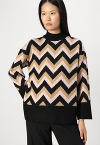 Pleteni pulover z obliko chevron v črni, bež in roza barvi, z visokim ovratnikom in ohlapnim krojem, izdelan iz mehkega teksturiranega materiala.
