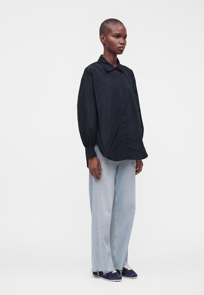Personne debout portant une chemise noire ample à boutons, un jean bleu clair à jambes larges avec des ourlets effilochés et des baskets bleu foncé sur un fond uni.