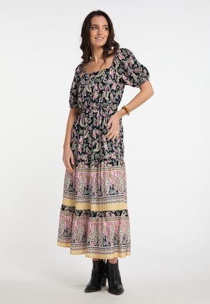 Frau trägt ein langes schwarzes, pinkes und beiges Blumen- und Paisley-Kleid mit Puffärmeln und schwarzen Stiefeletten, steht lächelnd.