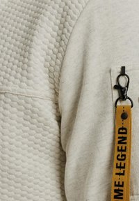 Sweatshirt beige clair avec un motif texturé en nid d'abeille, des coutures visibles, et une poche zippée sur le côté avec un clip noir et une sangle jaune.