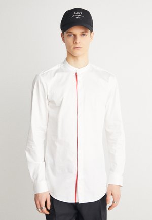Camicia bianca a maniche lunghe con colletto alla cinese, caratterizzata da una dettagliata patta dei bottoni rossa. La camicia è realizzata in tessuto liscio con un design aderente.