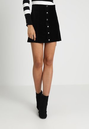 Kvinde iført en sort mini-nederdel med knapper foran, sorte ankelstøvler og en sort-hvid stribet langærmet top.