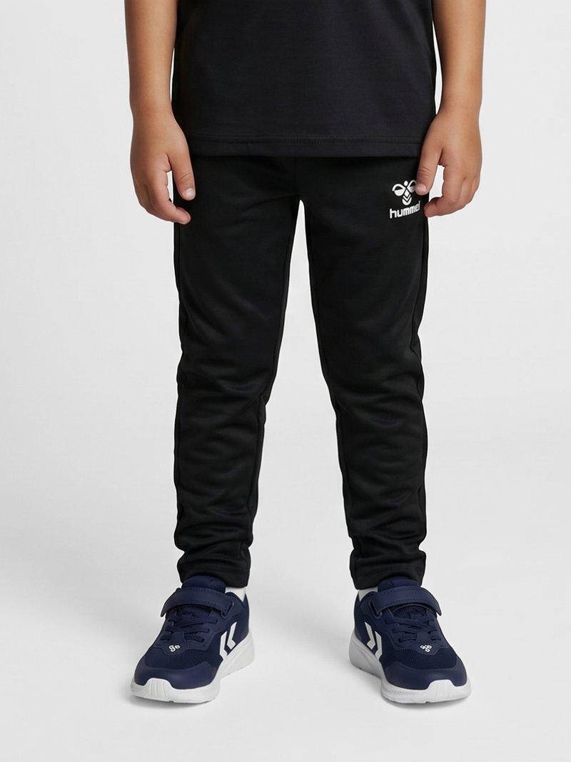 Barn iført sorte Hummel joggingbukser og marineblå sneakers står mod en lysegrå baggrund.