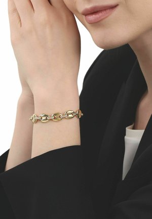 Primo piano di una persona che indossa un braccialetto d'oro con maglie ovali, le mani che sfiorano delicatamente il viso, vestita con abiti bianco e nero.