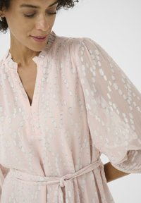Vestido ligero de color rosa con un patrón de lentejuelas plateadas, escote en pico, mangas abullonadas plisadas y un cinturón de lazo en la cintura. Tejido transparente y texturizado.