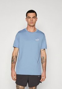 Camiseta de algodón azul claro con cuello redondo, mangas cortas y un pequeño logo blanco de Puma en el pecho. Combinada con pantalones cortos negros.