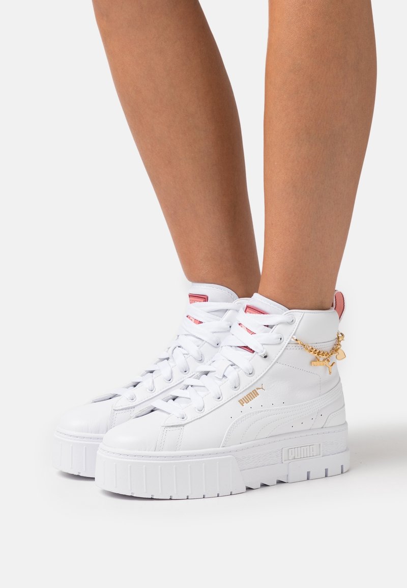 Baskets montantes blanches avec semelles épaisses, accents roses, logo Puma doré et détail chaîne dorée portées sur jambes nues.