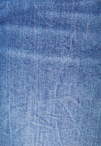 Tejido de mezclilla en un lavado azul medio, con una textura sutil que muestra patrones de tejido visibles y ligeras variaciones en el tono del color.