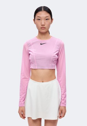 Kvinde med sort hår samlet på ryggen bærer en lyserød crop top med lange ærmer og en hvid nederdel, stående mod en ensartet lys baggrund.