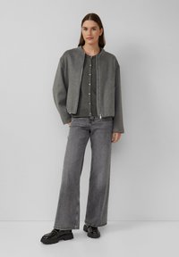 Grijze zip-up bomberjack met een ronde hals, gecombineerd met een grijze button-up shirt en wijde grijze jeans. Model draagt zwarte glanzende schoenen.