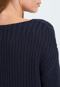 Mėlyna nertinė megztinė su plačiu, raštuotu kaklu ir tekstūrine, laisva struktūra. Dizainas rodo sklandų perėjimą tarp megztų raštų.