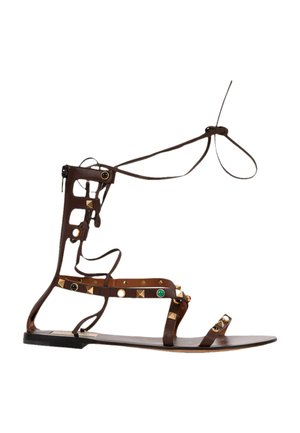 Bruine platte gladiatorensandaal met enkelriempjes, gouden studs en kleurrijke steenversieringen, met ritssluiting aan de achterkant.