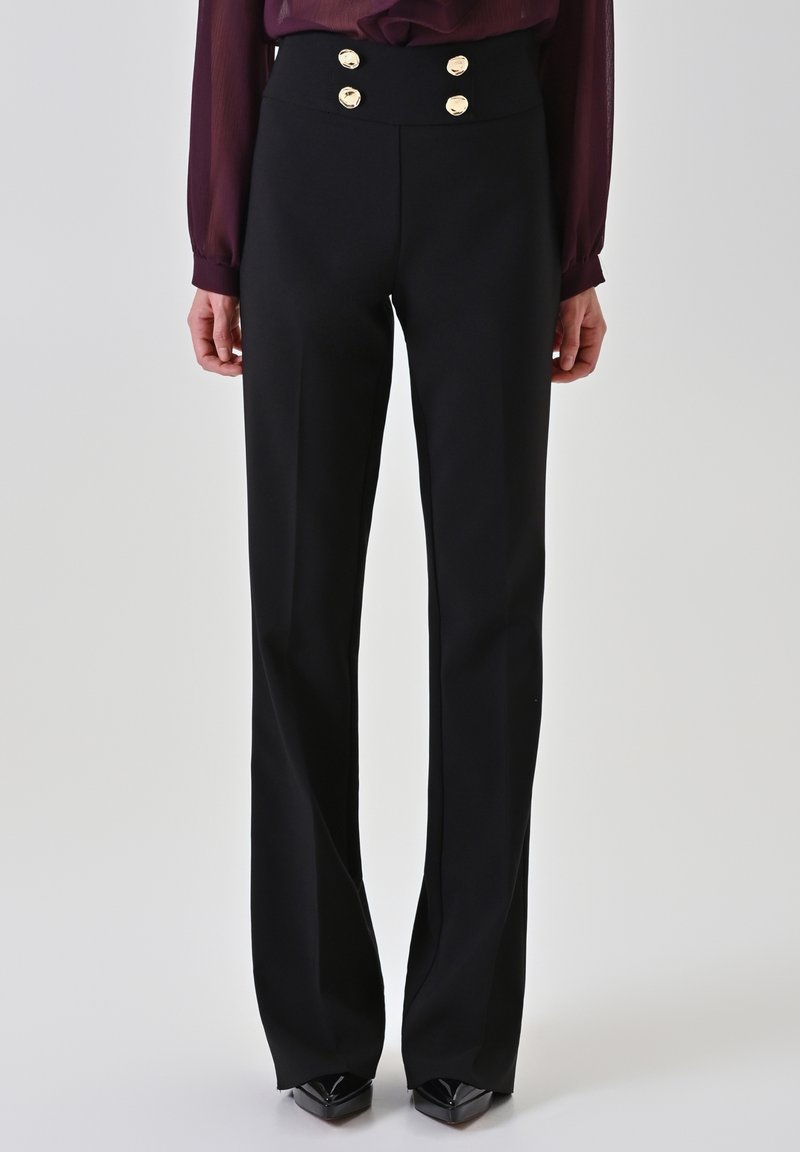 Vrouw draagt een zwarte broek met hoge taille en vier gouden knopen aan de taille, gecombineerd met een donkerpaarse blouse met lange mouwen en zwarte schoenen.