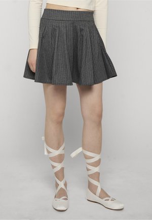 Mujer vistiendo una falda plisada de rayas grises y zapatos planos de ballet blancos con cintas cruzadas en los tobillos, de pie contra un fondo liso.