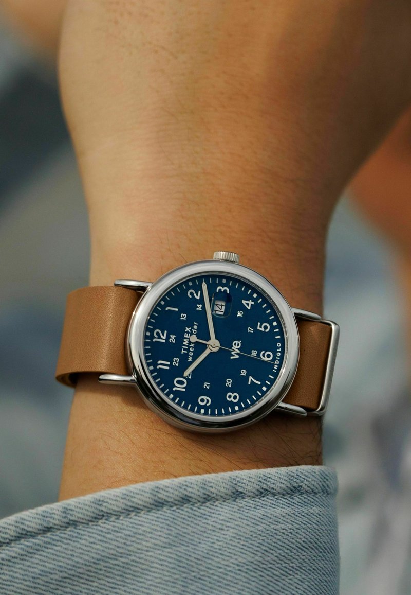 Montre Timex avec un cadran bleu, des chiffres blancs et un affichage de la date. Boîtier en métal et bracelet en cuir marron. Design décontracté avec une visibilité claire.