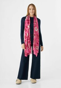 Écharpe florale rose avec des bords frangés, en tissu léger, ornée de motifs floraux blancs et rouges ; portée avec un haut à manches longues bleu marine et un jean large.