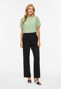 Blouse en dentelle vert clair à manches courtes, associée à un pantalon noir taille haute et des talons ouverts noirs. Design simple et structuré.