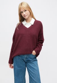 Bordový oversize sveter s výstrihom do V a rebrovanými manžetami, nosený cez bielu košeľu s golierom. Vyznačuje sa mäkkou, textúrovanou látkou.