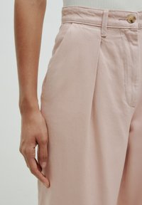 Lichtroze, hooggesneden broek met een vlakke voorkant, zijzakken en een enkele knoopsluiting. De stof heeft een gladde, licht gestructureerde afwerking.