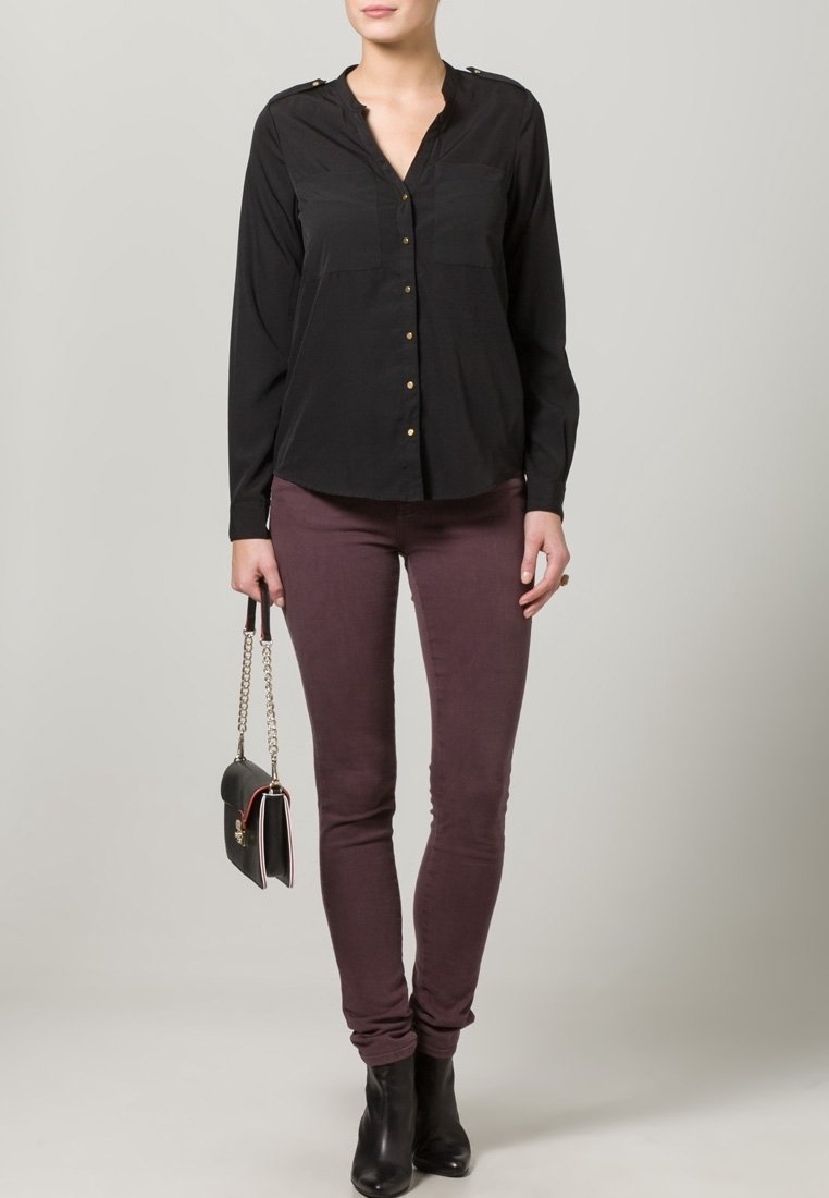 Camicia nera a maniche lunghe con bottoni e dettagli dorati, abbinata a pantaloni bordeaux aderenti e stivaletti neri, mentre tiene una piccola borsa nera.