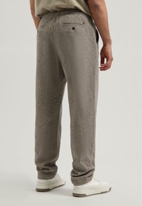 Man met een losse bruine geruite broek en witte sneakers, staand met zijn rug naar de camera op een effen achtergrond.