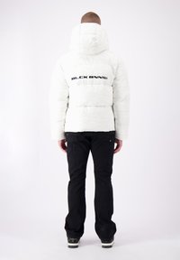 Witte pufferjack met capuchon, met opvallende zwarte tekst "BLCK BNNS" op de achterkant. Gecombineerd met zwarte broek en schoenen, wat een casual stijl laat zien.