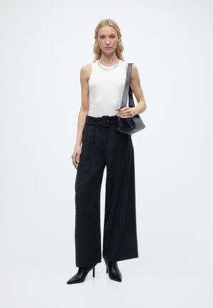 Γυναίκα που φοράει λευκό αμάνικο τοπ, μαύρο παντελόνι με φαρδιές μπατζάκια και ψηλή μέση με ζώνη, μαύρες μπότες με τακούνι, και κρατάει μαύρη τσάντα ώμου.