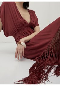 Maroon dress with a deep V-neck, pleated bodice, and a flowing silhouette. Features side fringe detailing and an oversized cuff bracelet.