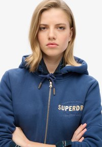 Sudadera con cremallera azul y capucha con cordón, bordado en dorado que dice "SUPERDRY" y "New York City", con tejido texturizado y un ajuste relajado.