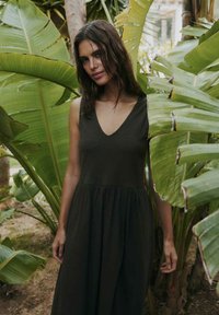 Woman with long dark hair wearing a sleeveless black dress stands among large green tropical leaves outdoors.